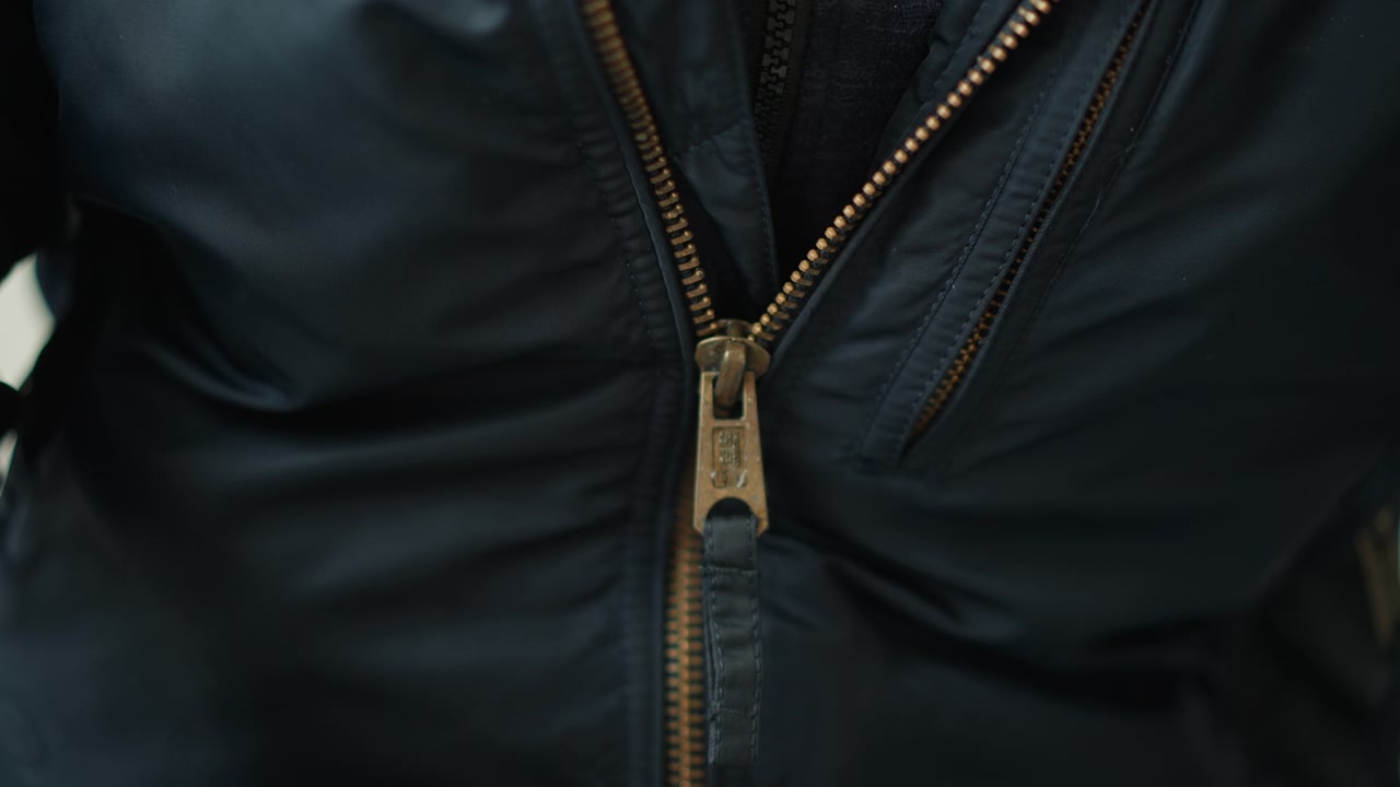 Nahaufnahme einer Person, die eine dunkelblaue Jacke trägt, deren Reißverschluss teilweise geöffnet ist und einen Teil des darunter liegenden Kleidungsstücks enthüllt. Der Fokus liegt auf der Textur und den Details des Stoffes und des messingfarbenen Reißverschlusses. Weiche Beleuchtung.