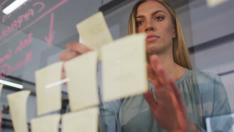 caucasian businesswoman brainstorming using memo notes on glass wall in office and smiling