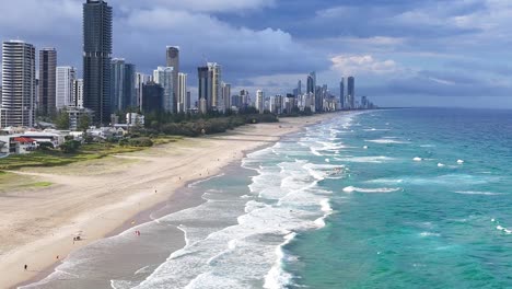 aerial view of a vibrant city skyline with waves crashing on a sandy beach under a partly cloudy sky.