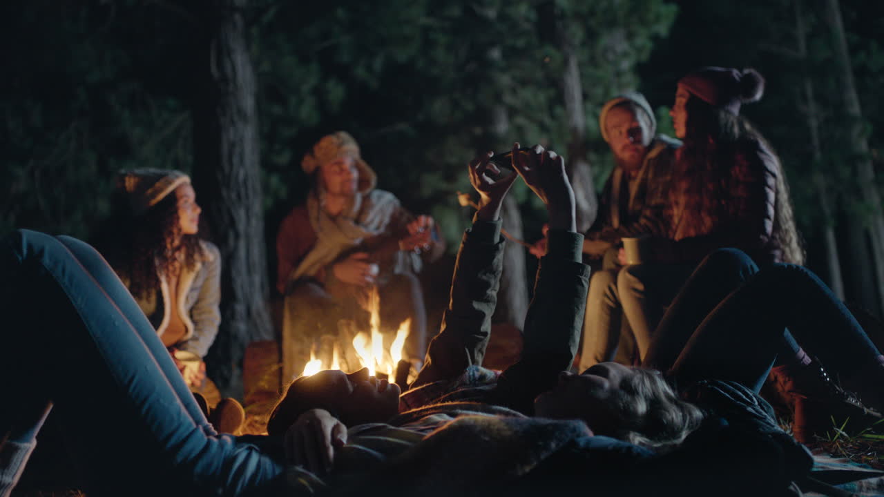 스마트폰을 사용하는 캠핑 친구들은 밤에 숲 속의 모닥불 옆에 누워 휴대폰 카메라 플래시 공유 모험과 함께 사진을 찍습니다.