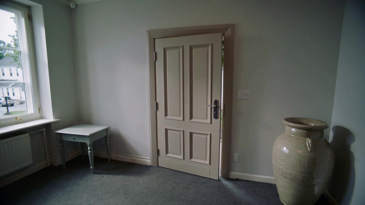 Porta de madeira com painéis balançando empurrada pela brisa externa, fechando-se em sala interior para bloquear a rua.