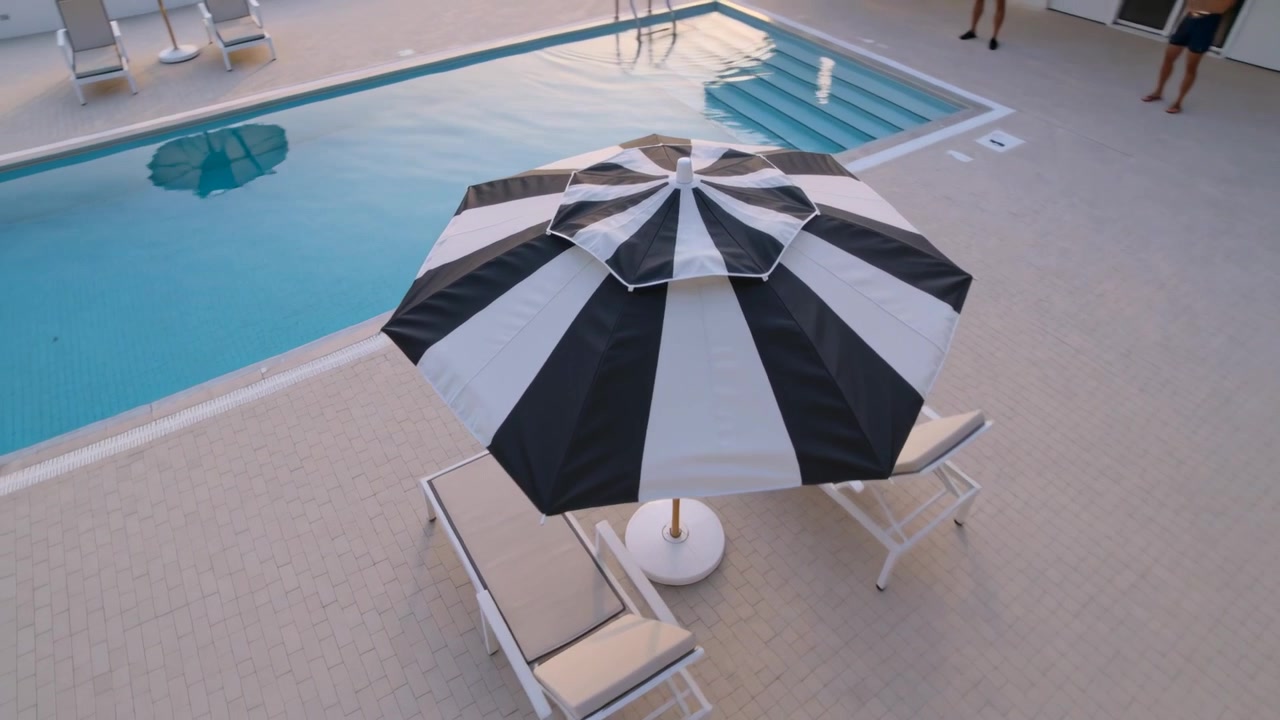 Inizio ripresa con telecamera che si muove sopra la piscina, centrando un ombrellone a strisce e lettini, uomini in piedi in pantaloncini.