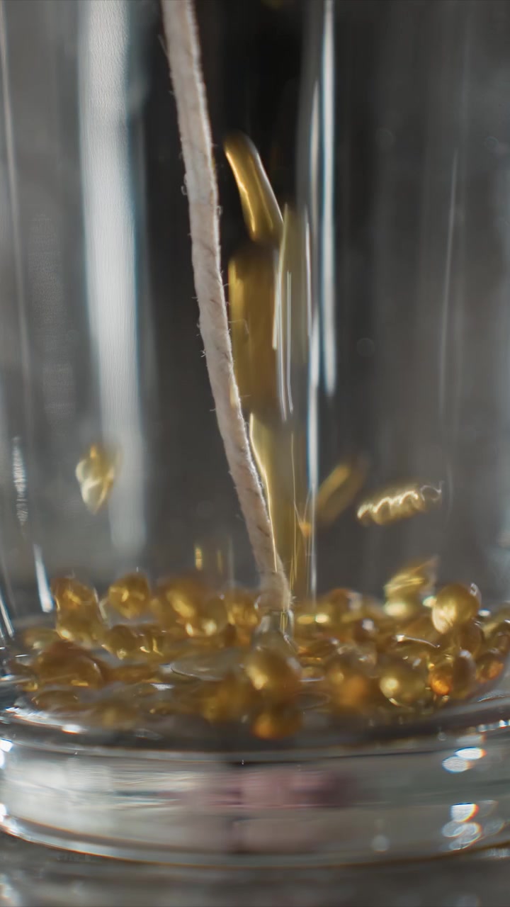 Primo piano di goccioline di liquido dorato che cadono in un contenitore di vetro con illuminazione soffusa. Il flusso continuo crea increspature e si accumula alla base, evidenziando i dettagli della texture in primo piano.
