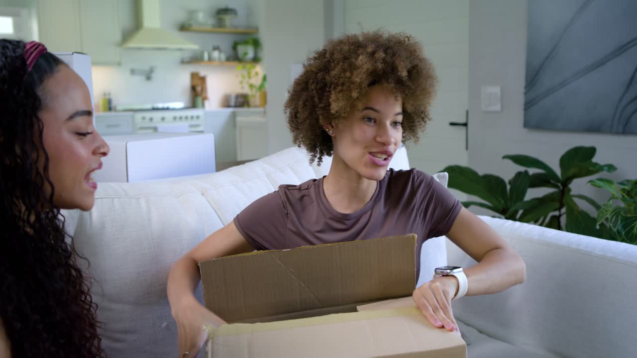 Two African American women opening sealed box on couch, revealing bubble wrap, unwrapping appliance