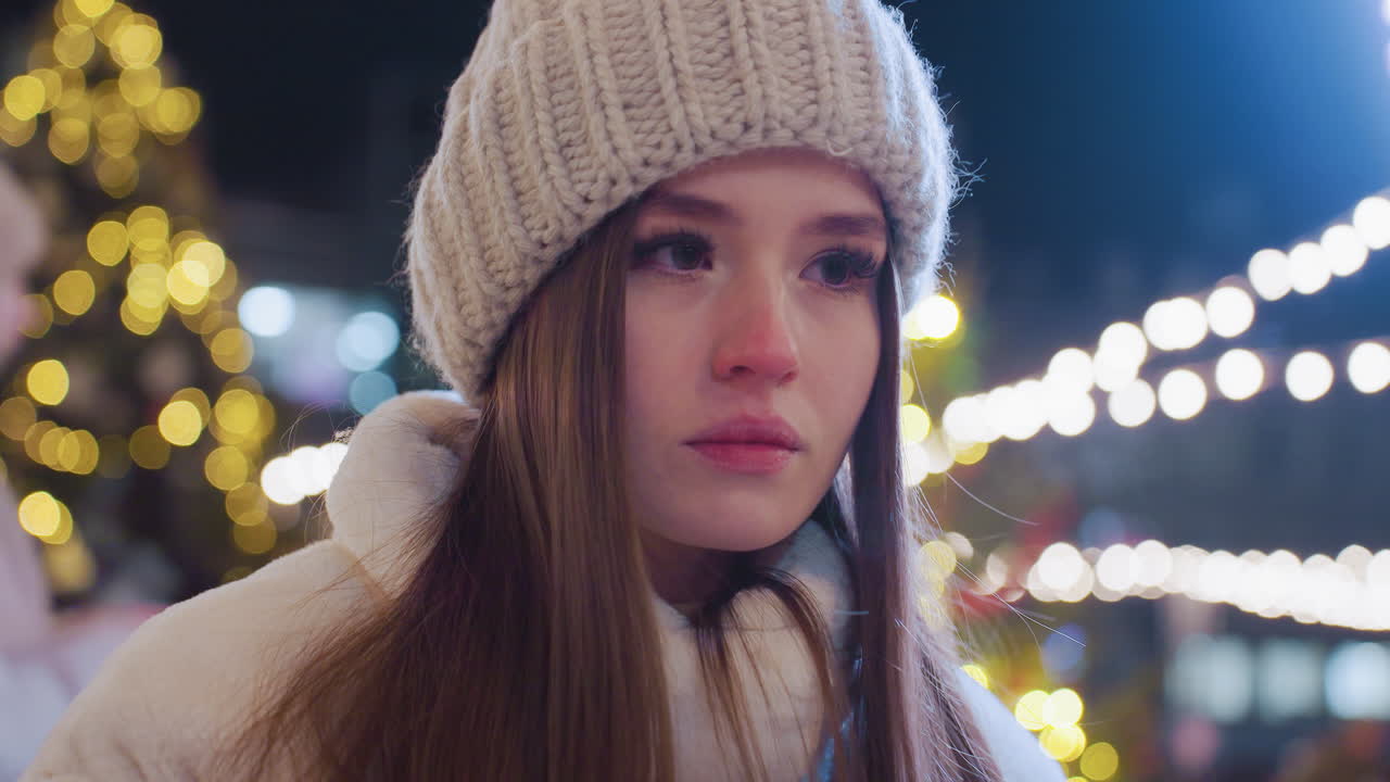 Young woman in warm winter clothing sits outdoors at night, appearing sad and contemplative, city lights glow around her as people skate, socialize, and celebrate