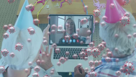 animación de células de covid-19 flotando sobre una pareja mayor con sombreros de fiesta usando una computadora portátil en una llamada de video