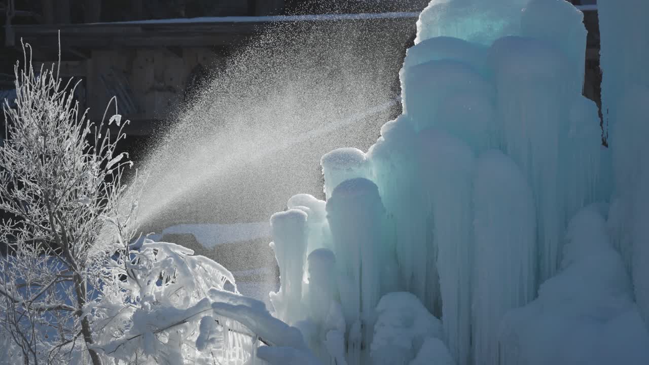 Water jets, backlit by the morning sun, spray over a towering blue ice mound in a winter forest scene.