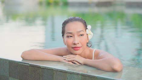 Portrait-of-a-young-woman-with-flower-in-hair-leaning-on-the-edge-of-a-swimming-pool-in-a-Tropical-spa-resort-in-Hawaii,-face-close-up