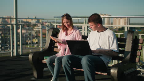 two young adults studying outdoors, male on laptop while female reads book and points out details, set against an urban landscape with modern buildings and lush greenery