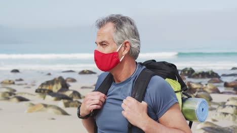 Portrait-of-senior-hiker-man-wearing-face-mask-with-backpack-hiking-on-the-beach.