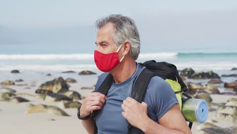 retrato de un hombre excursionista mayor con una máscara facial y una mochila caminando por la playa.