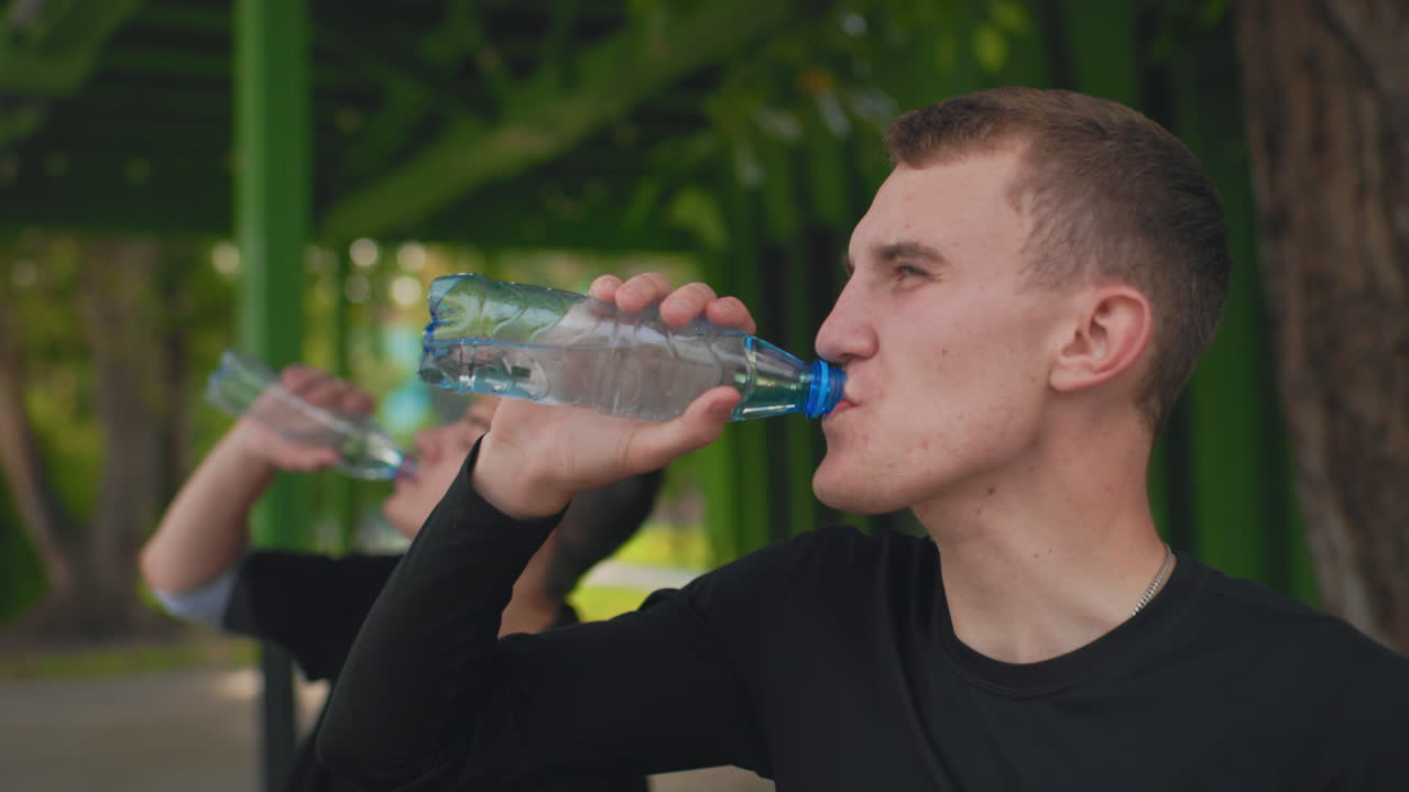Молодой человек пьет воду после тренировки под зеленым навесом на скамейке в парке; на заднем плане — друг. Случайная летняя сцена, показывающая пот, глубокое дыхание, непринужденный разговор, глоток из бутылки.
