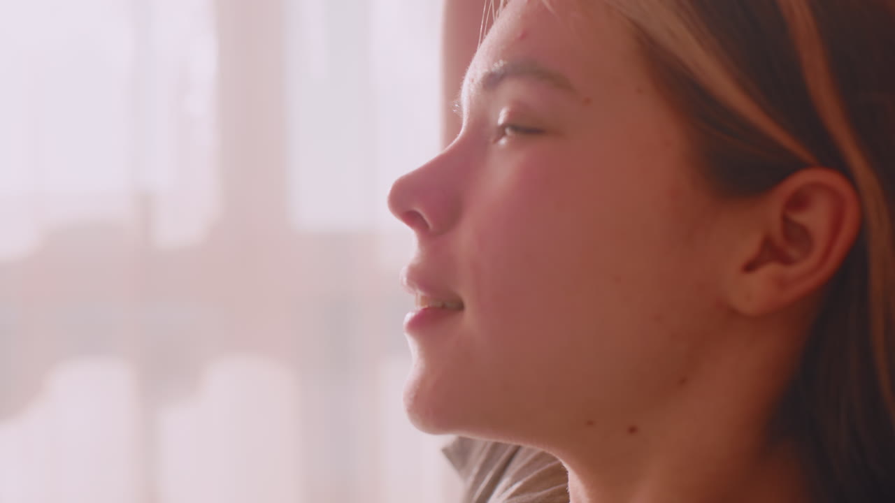 Close up of young woman raising arms and stretching with eyes closed in soft morning glow, enjoying peaceful start to day with relaxed face and gentle expression surrounded by translucent curtains