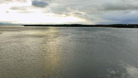 Vast waterscape reflecting overcast sky on dull daytime. Few cars move by the highway on the waterfront. Aerial view.