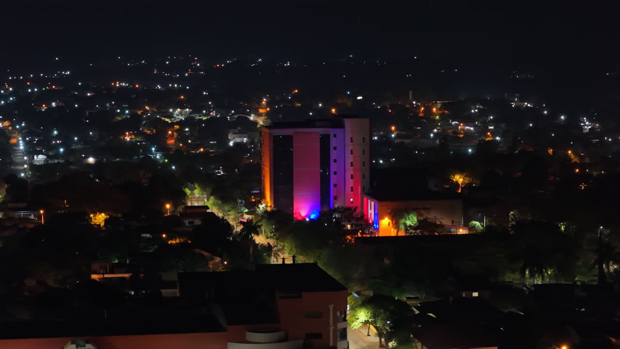 Vista aérea de um edifício de apartamentos colorido de média altura e da paisagem urbana noturna em Villarrica, Paraguai.