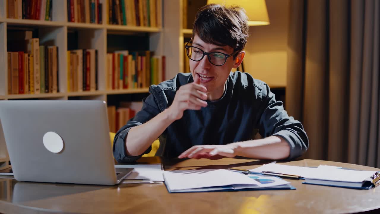A young man gestures enthusiastically in a video call, seated at a round table with a laptop