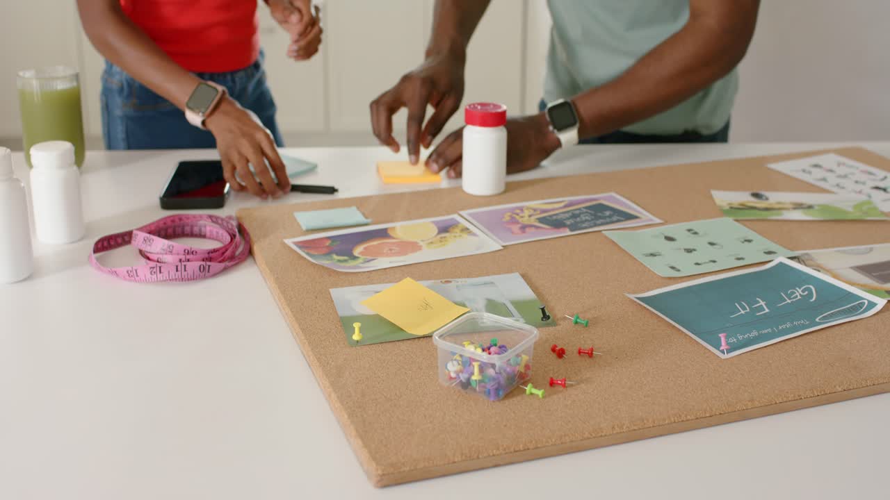 African American pair arranging sticky notes on board at kitchen counter, man holding jar planning