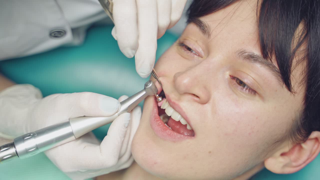Young woman receives professional teeth cleaning in cosmetic dentistry clinic. Patient's face and stomatologist working with a tool which has small brush in dentist office. Close-up