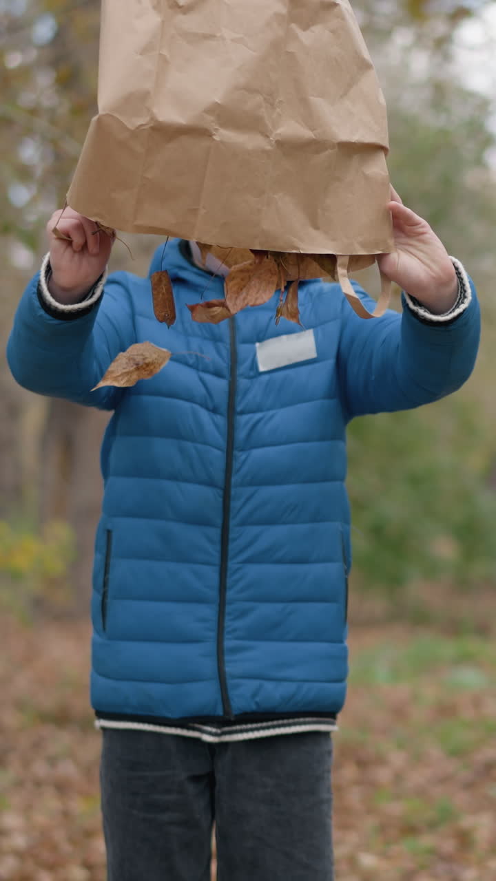 어린 아이가 건조한 잎을 따라 고, 기게 종이 봉투를 거꾸로 돌리고, 땅에 건조 한 잎을 부어, 활기찬 초록색과 시골 울타리에 둘러싸여 가을 설정