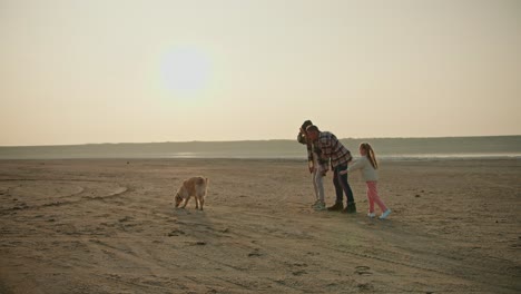 Happy-family-playing-with-their-pet-dog-outside-the-city-while-relaxing-in-the-summer-evening.-A-middle-aged-brunette-man-in-a-checkered-shirt-and-his-wife-and-little-daughter-play-with-their-large-cream-colored-dog-on-a-deserted-seashore-during-their-vacation-outside-the-city-in-the-evening-in-summ