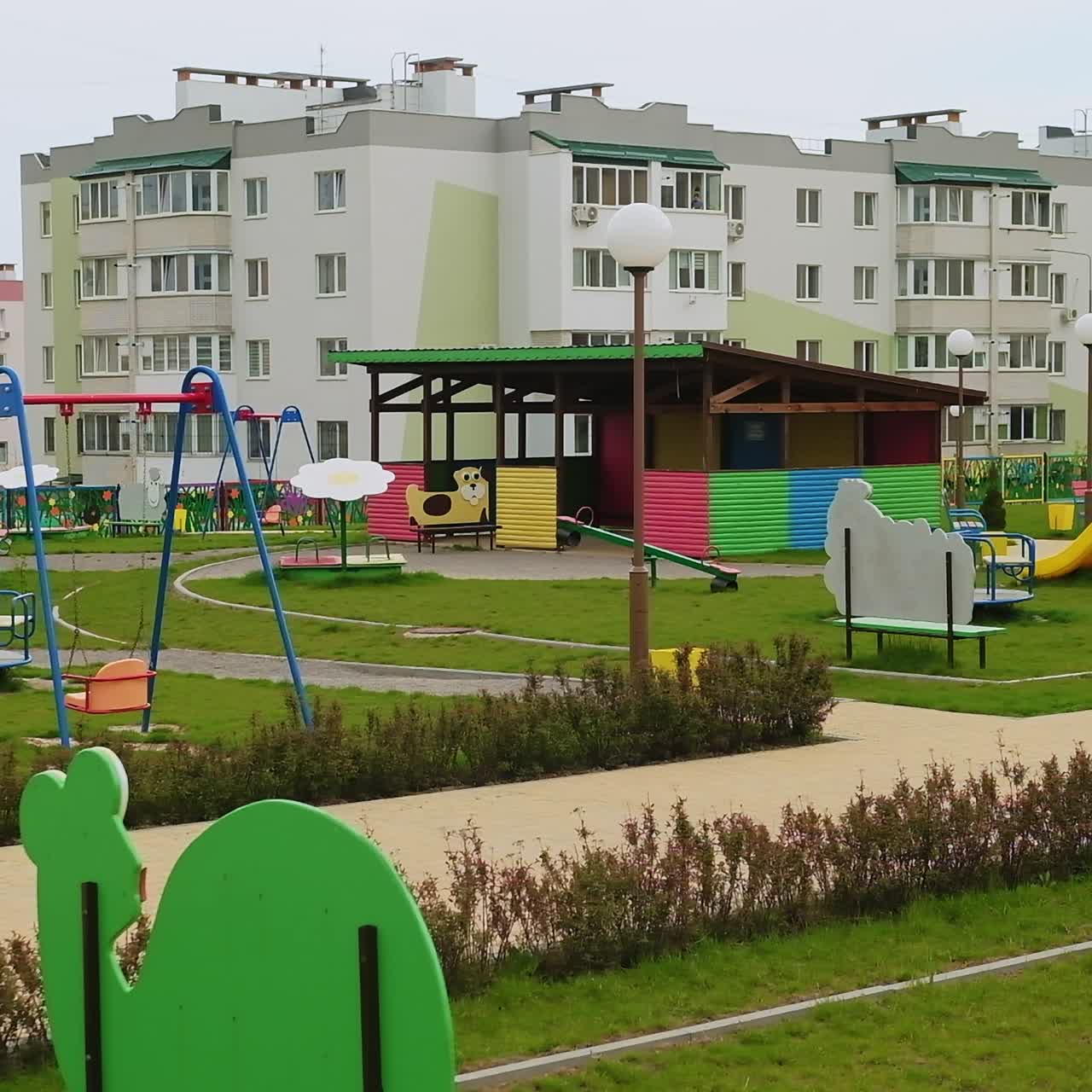 Well-kept territory of newly-built kindergarten. Big gazebos and teeterboards on the green lawn at the backdrop of new houses