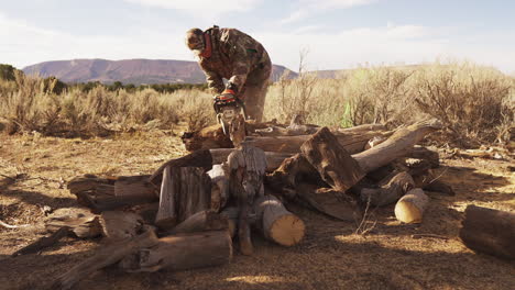 late afternoon, golden hour, a man in camp gear approaches a pile of firewood with a chainsaw and starts cutting