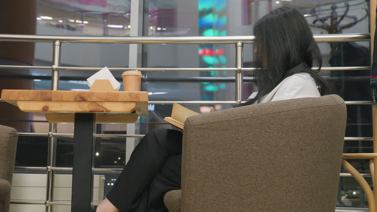Side view of student seated in restaurant reading book with tattoo on hand while flipping to new page, colorful lights blinking through window in background