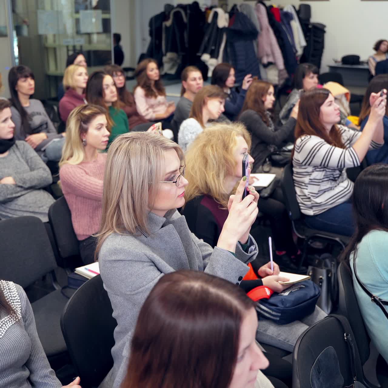 Crowded auditorium indoors. Women sitting on the chairs in the hall and taking photographs on their cell phones during seminar.