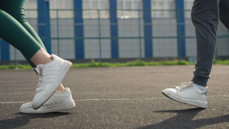 leg view of two women jogging on sports court, one in green joggers and one in ash joggers with white sneakers, blurred background featuring bar fence and people seated outdoors