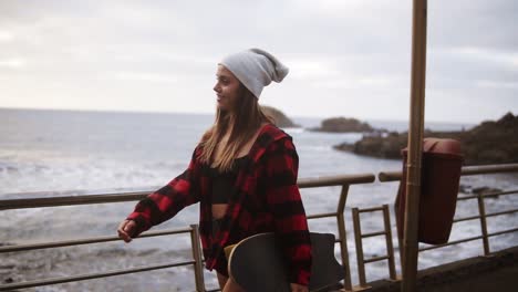 Nahaufnahmen-Sportliche-Frau-Mit-Hut,-Karierter-Mantel,-Die-An-Einem-Bewölkten-Tag-Bei-Sonnenuntergang-Die-Zeit-Am-Meer-Genießt,-Freier-Geht-Und-Ein-Skateboard-Hält