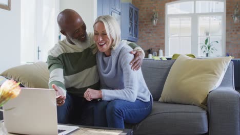 Happy-senior-diverse-couple-in-living-room-sitting-on-sofa,-using-laptop,-making-video-call