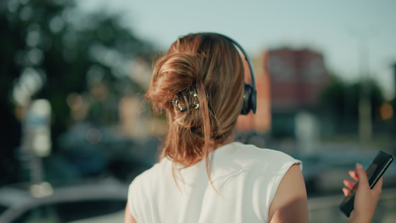 Close up of white lady with hair clip enjoying music through headphones while facing blur urban background with parked cars, giving relaxed vibe of personal enjoyment in city environment