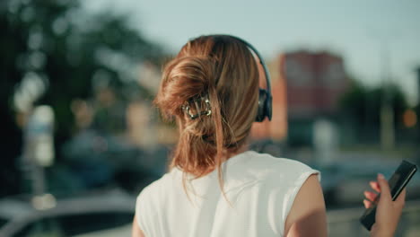 close up of white lady with hair clip enjoying music through headphones while facing blur urban background with parked cars, giving relaxed vibe of personal enjoyment in city environment