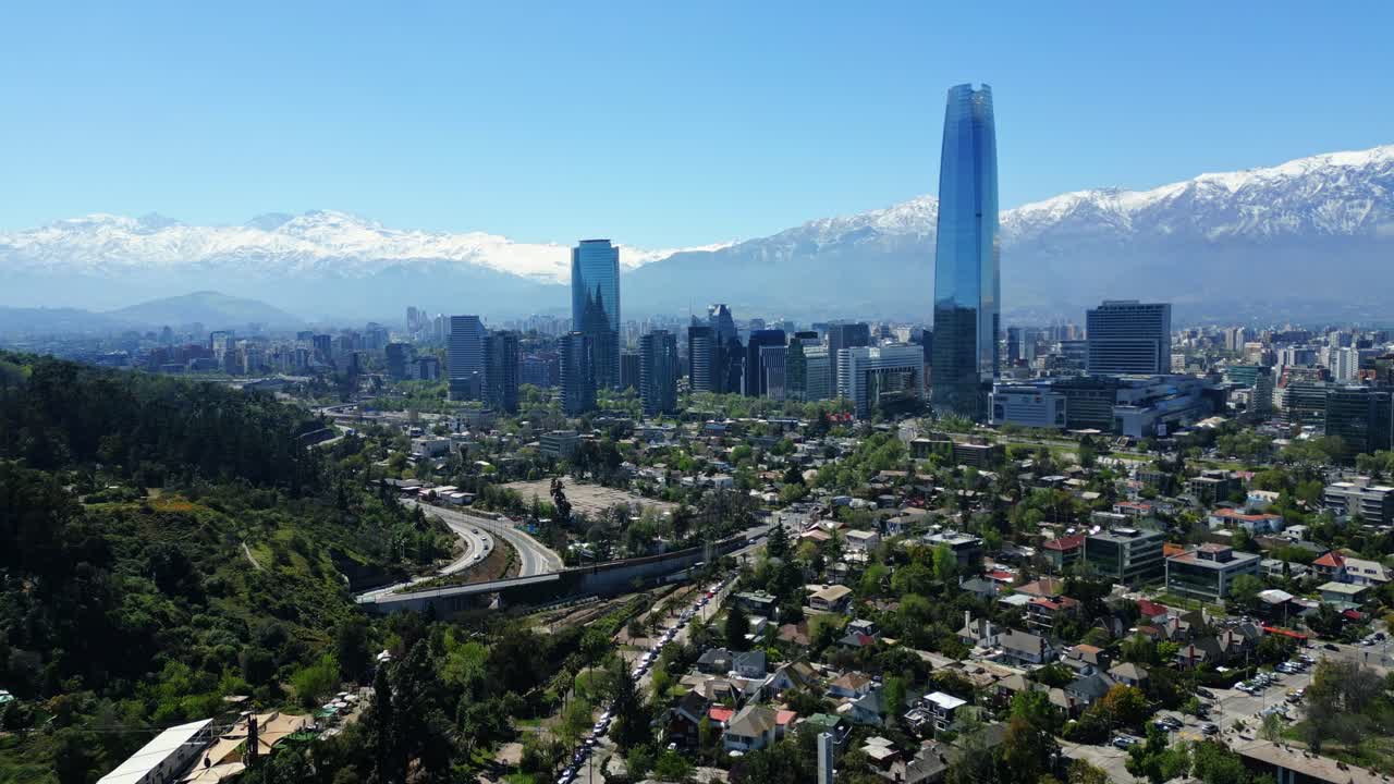 Drone aerial forward ascending over treelined hills, rooftops and highways toward the glass Costanera tower and snowcapped Andes in clear daytime light. Santiago, Chile