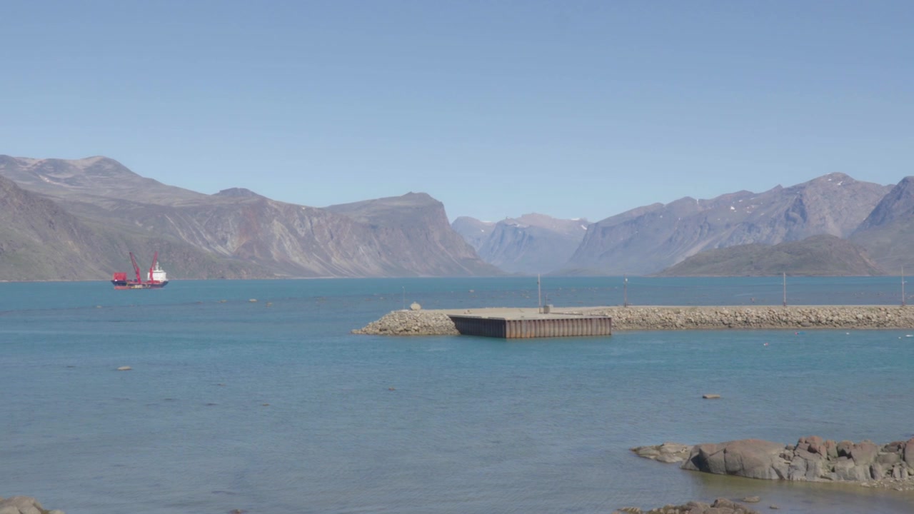 Panoramica fluida verso sinistra attraverso l'infrastruttura della strada rialzata in ghiaia e le aree di allestimento industriale per rivelare operazioni attive di trasporto marittimo nel porto di Pangnirtung.