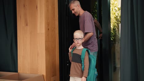 a dad with gray hair in a purple t-shirt came with his son with white hair in round glasses and a green backpack to a club for preparing children for school where they were met by a woman teacher in a white shirt