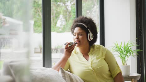 Video-of-happy-plus-size-african-american-woman-with-headphones-sitting-on-sofa-with-smartphone