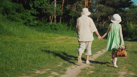 Grandmother-Is-Walking-With-Her-Granddaughter-In-The-Forest-Holding-Hands-The-Girl-Carries-A-Basket-