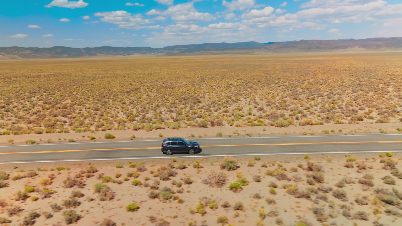 Cars going by the empty road in the sunny desert. Mountainous horizon line under blue cloudy sky at backdrop.