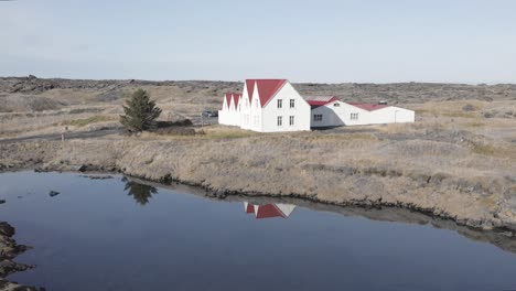 Aerial-of-idyllic-red-and-white-traditional-house-next-to-calm-lake-in-Iceland