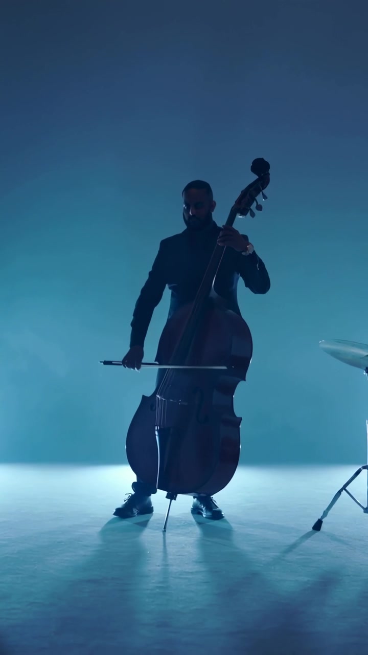 Video verticale: Bassista che si prepara suonando il contrabbasso con l'archetto su un palco illuminato di blu, giacca scura, batteria.