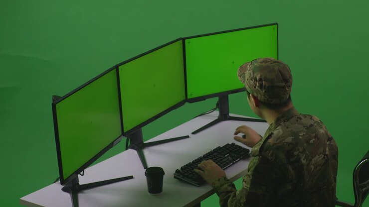 Sidebillede af asiatisk mandlig soldat, der tænker over noget og derefter løfter sin pegefinger, mens han arbejder med mockup af flere computerskærme i et green screen-studie.
