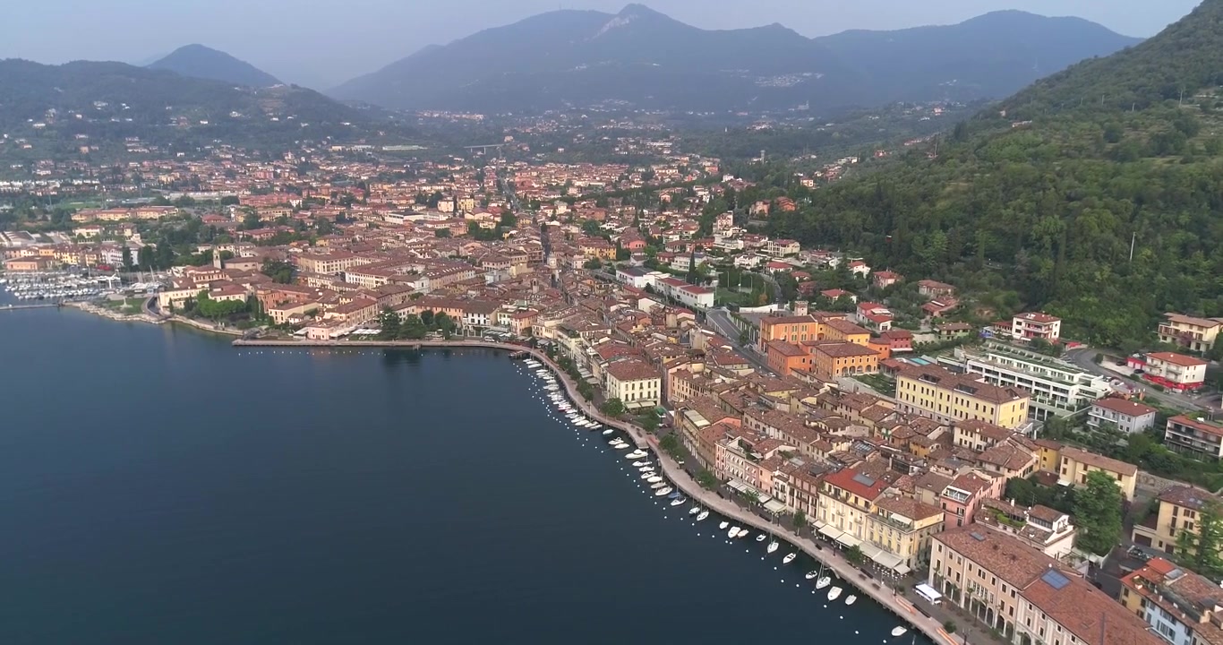Vista aerea con drone della città di Salò con fitti tetti in terracotta lungo la costa curva del Lago di Garda, con acqua calma e colline verdi circostanti sotto una luce diurna soffusa.