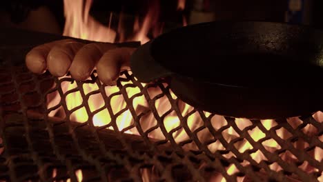 Hot-orange-camp-fire-flames-illuminate-sausages-and-pan-on-metal-grill