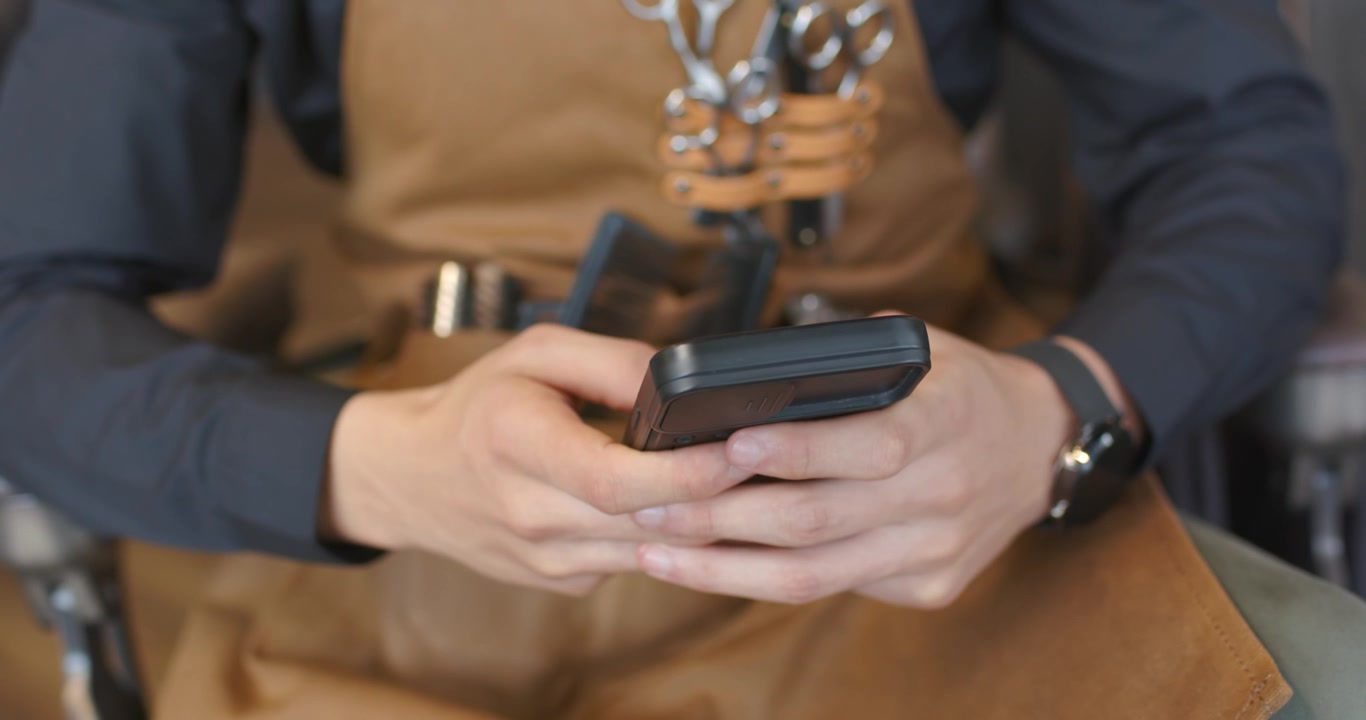 Barbiere adulto che controlla il telefono, toccando e scorrendo, in un negozio da barbiere, con grembiule e forbici
