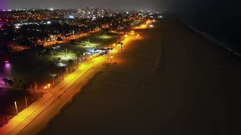 Luftflug-über-Den-Strand-Von-Santa-Monica-Bei-Nacht-Mit-Radweg,-Der-Von-Gelben-Lichtern-Beleuchtet-Wird
