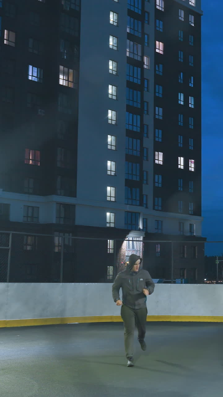 Soccer player kicks ball toward goalpost under bright urban lights, seen through entrance on outdoor field surrounded by tall residential buildings against a vibrant blue evening sky
