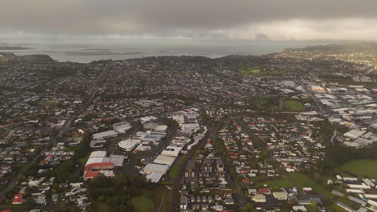 Wide aerial shot over Avondale suburb, Auckland, residential neighborhoods, commercial zone, harbour in distance under cloudy sky, suburbia. New Zealand. Drone pov