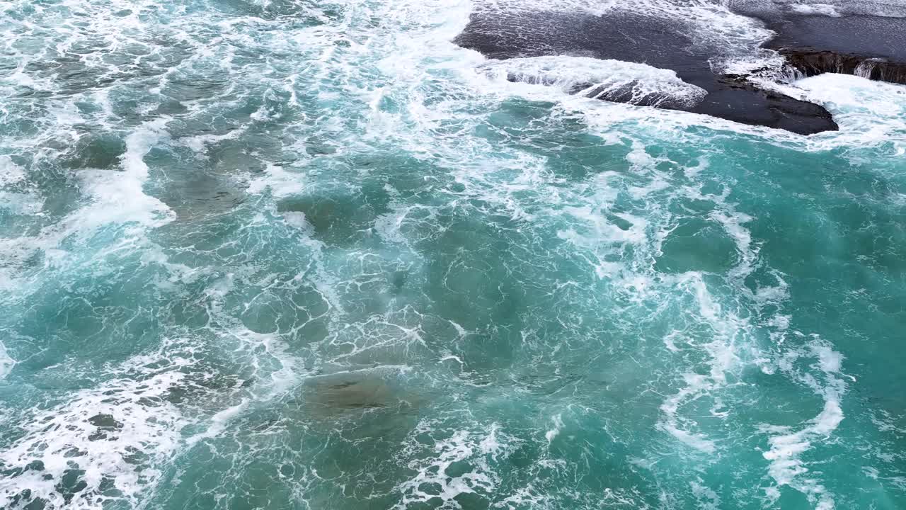 Overhead drone shot of turquoise waves breaking against rocks, natural daylight, dynamic water movement