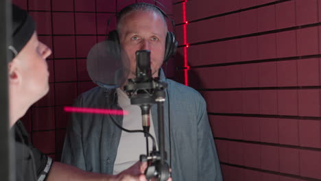 a man in a blue shirt and white inner shirt stands in a red soundproof room wearing headphones, as another man in a black shirt and cap adjusts the microphone in front of him